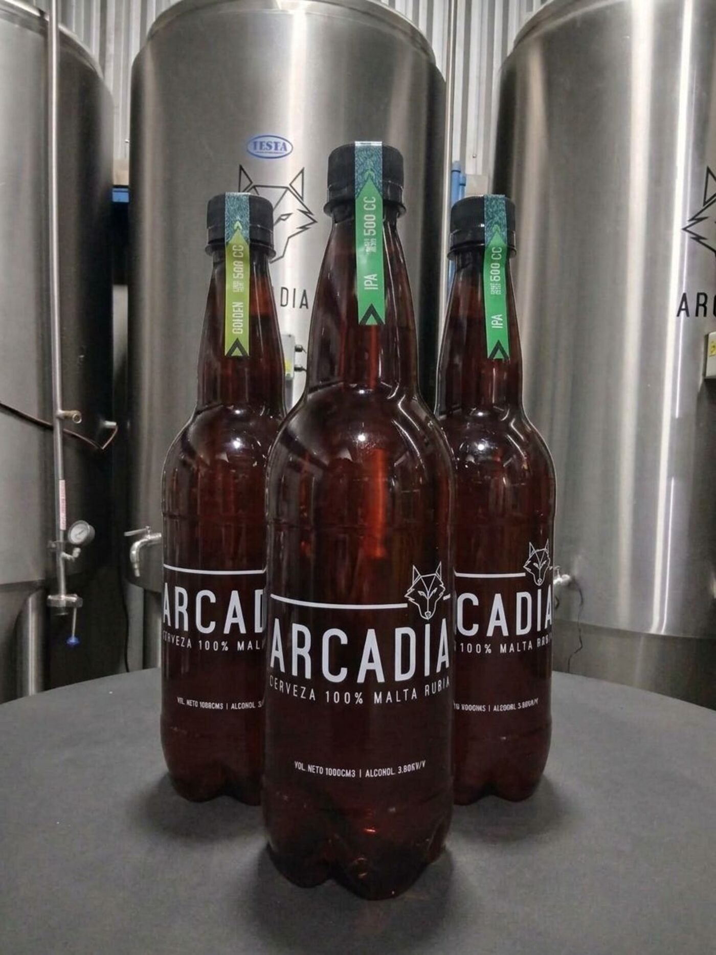Tres botellas de Arcadia frente a los fermentadores de acero de la fábrica